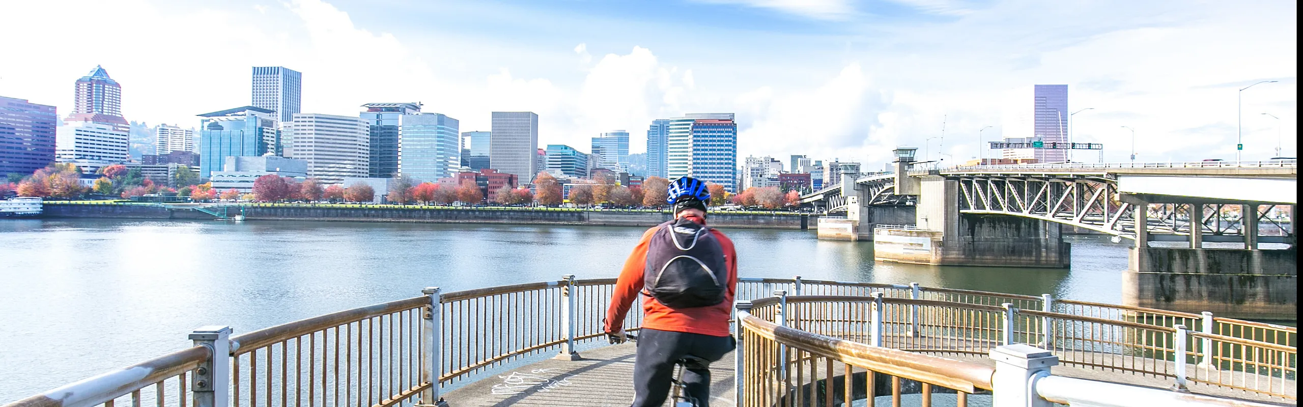 Vibrant cityscape view from a riverside bike path, featuring a cyclist riding towards a skyline of modern skyscrapers and a scenic bridge.
