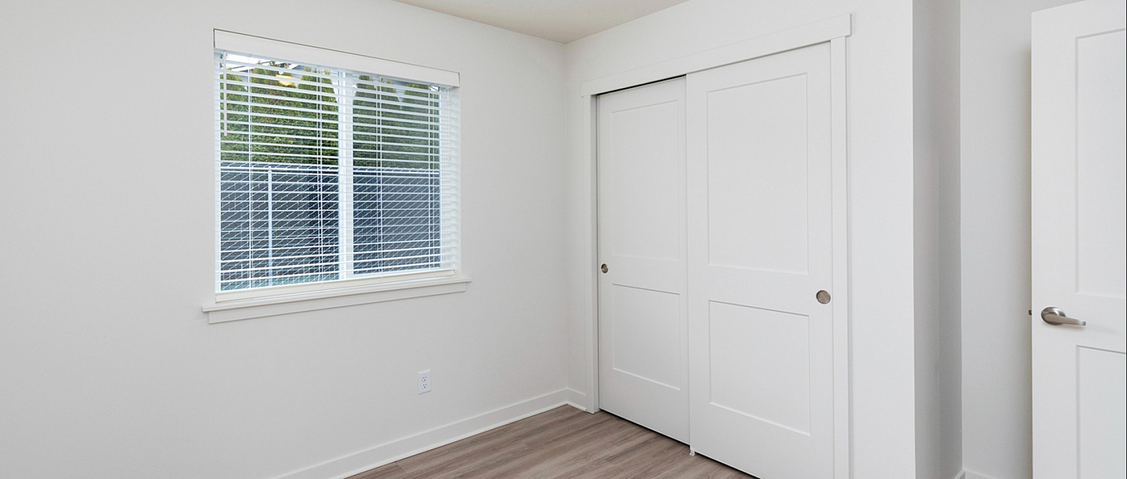 Bright and airy bedroom with sleek wooden flooring, ample natural light from a large window, and spacious closet with sliding doors.