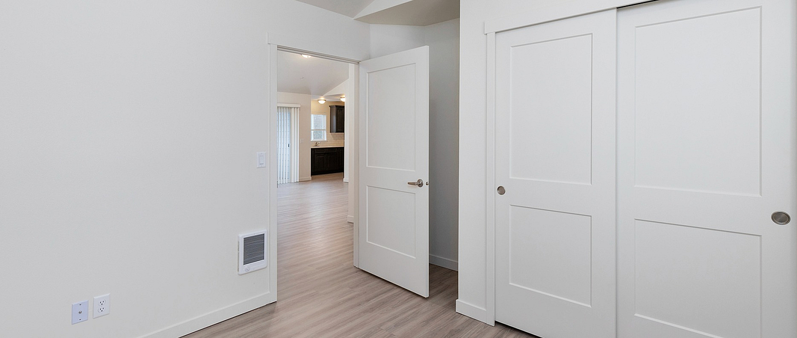 Bright and spacious room featuring elegant wood flooring, sleek white walls, and modern sliding closet doors, leading to an inviting open-concept living area.