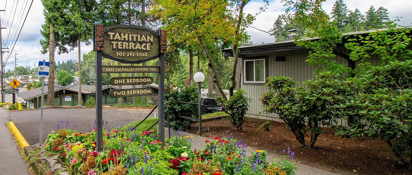 Inviting entry to Tahitian Terrace apartments with vibrant flower beds, surrounded by lush greenery and serene neighborhood views.