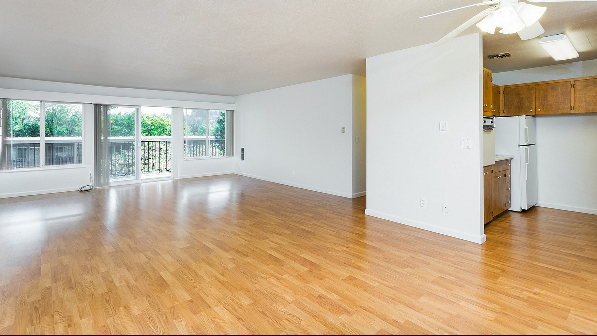 Bright, spacious open-plan living area with hardwood floors and large windows showcasing lush greenery, adjacent to a cozy kitchen.