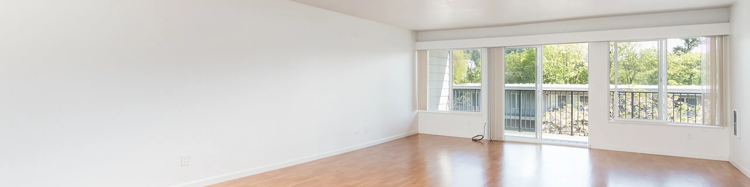 Bright, spacious living room with sleek hardwood flooring, large windows offering abundant natural light, and a view of lush greenery.