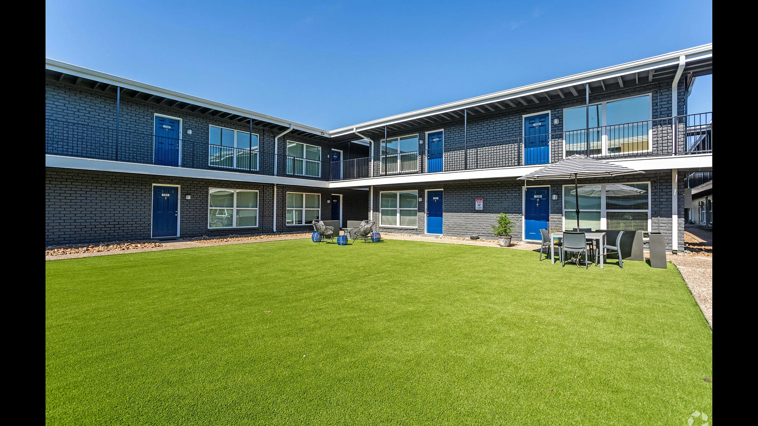 Modern apartment complex featuring a lush green courtyard, stylish blue doors, and inviting outdoor seating area under a chic umbrella.