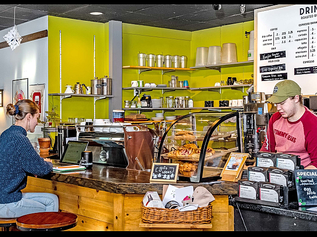 Cozy coffee shop interior with vibrant green walls, displaying a welcoming ambiance. A customer sits at the counter with a laptop, as a barista prepares drinks. The counter showcases a variety of pastries and an array of coffee options on the menu, enhancing the inviting atmosphere.