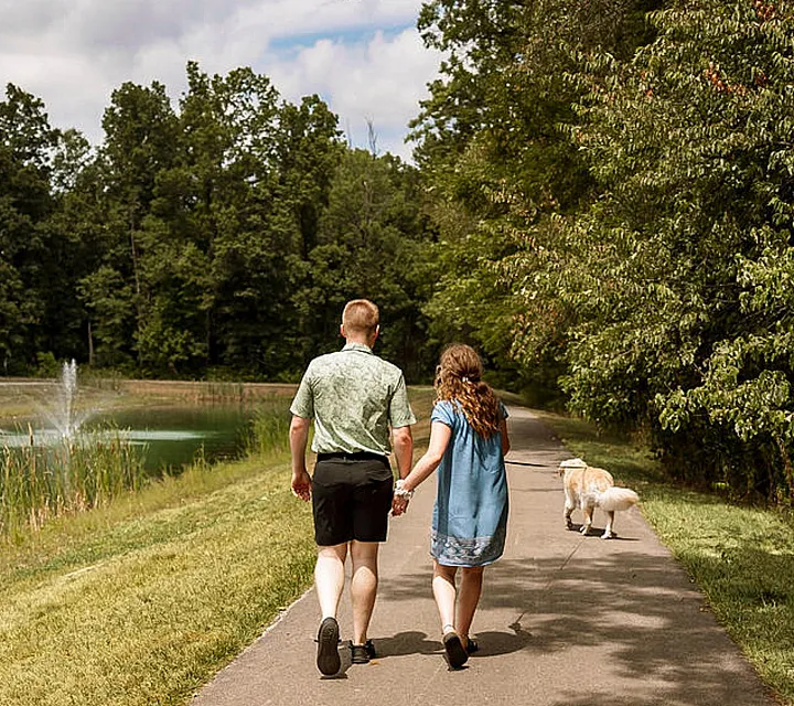 A couple strolls hand-in-hand down a scenic park path surrounded by lush greenery and a tranquil pond.