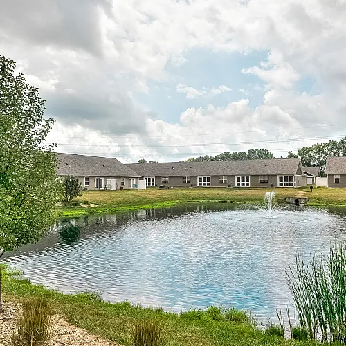 Picturesque view of modern apartments by a tranquil pond with a fountain and lush greenery.