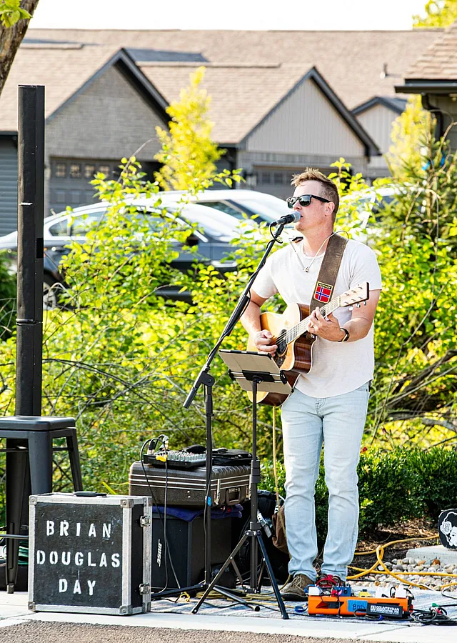 Vibrant outdoor performance featuring a musician playing guitar and singing, with houses and greenery in the background.