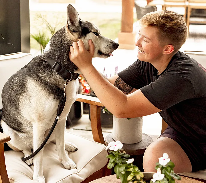 Cozy seating area with a smiling person gently holding their affectionate husky, surrounded by vibrant potted plants, offering warmth and comfort.