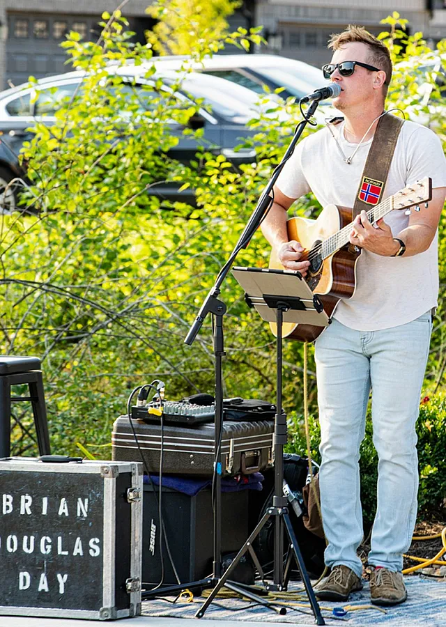 Outdoor music performance with a guitarist singing passionately on a sunny day, surrounded by lush greenery and quaint houses.