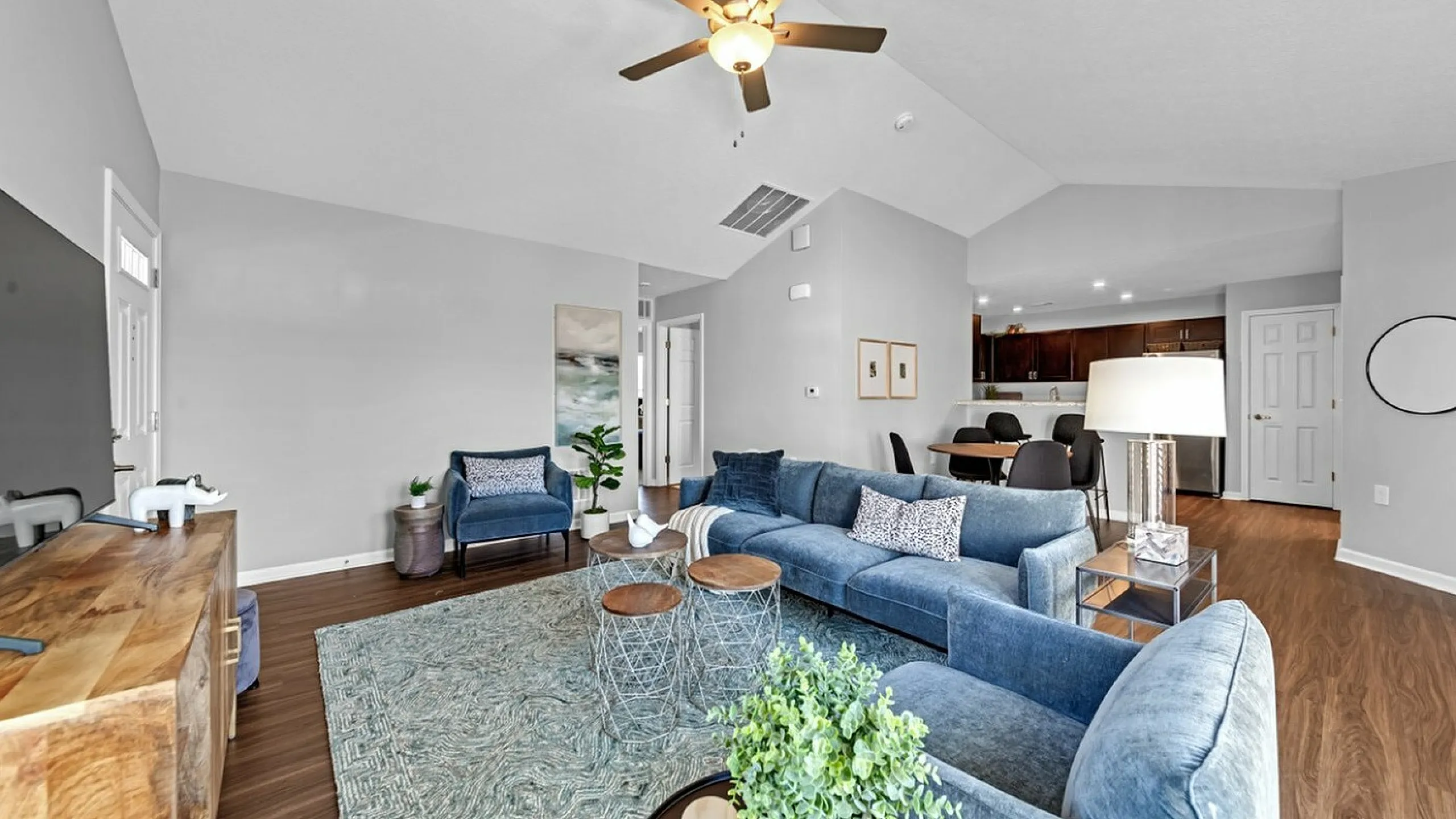 Inviting open-concept living room featuring plush blue furniture, elegant wood flooring, and a stylish ceiling fan, seamlessly flowing into a modern kitchen and dining area.