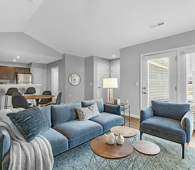 Bright, modern living room with plush blue seating, stylish accent tables, and an open view to a sleek kitchen area.