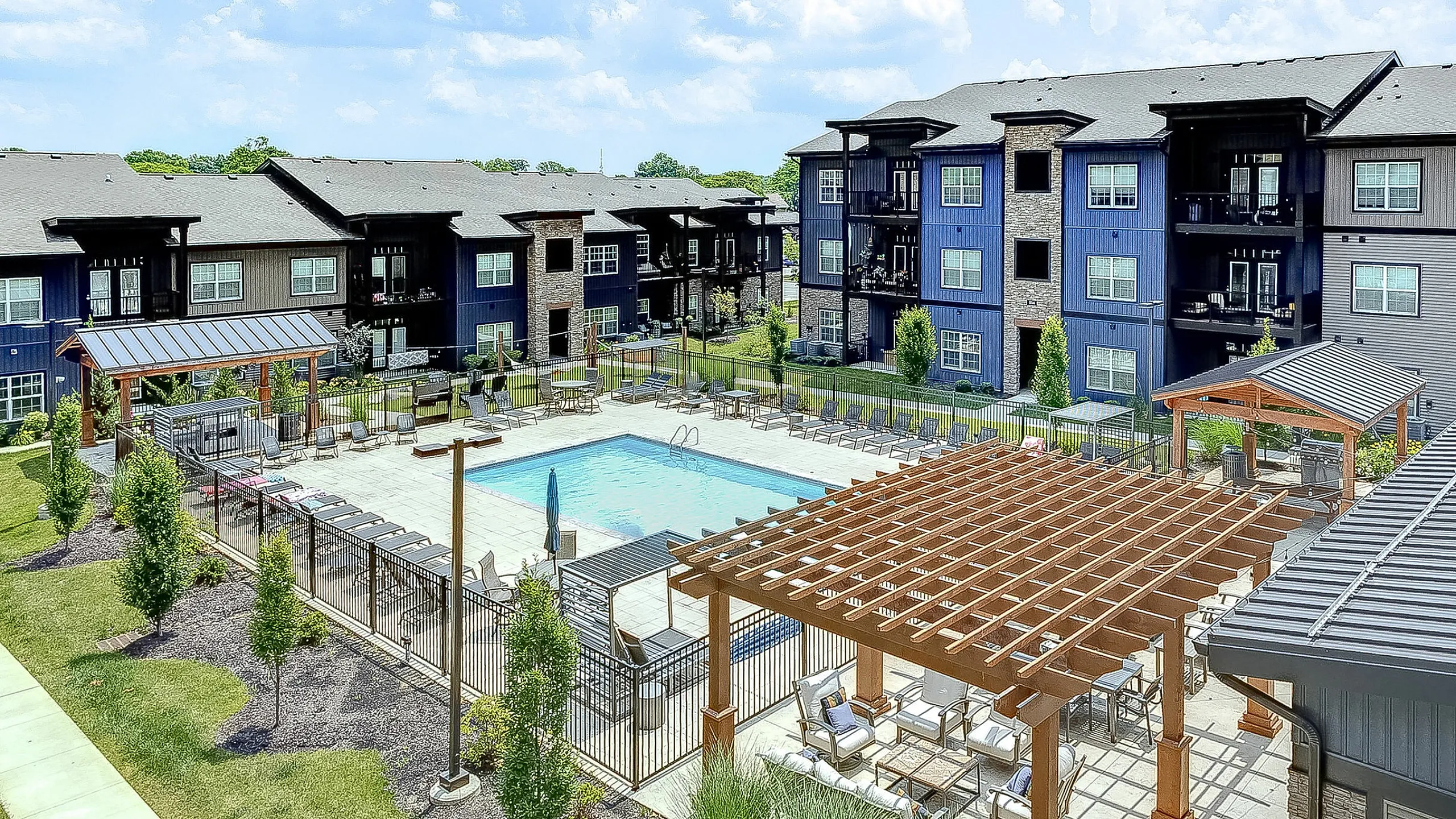 Charming residential complex with modern apartments, a serene swimming pool, and inviting outdoor seating areas under a wooden pergola, surrounded by lush greenery and bright blue skies.
