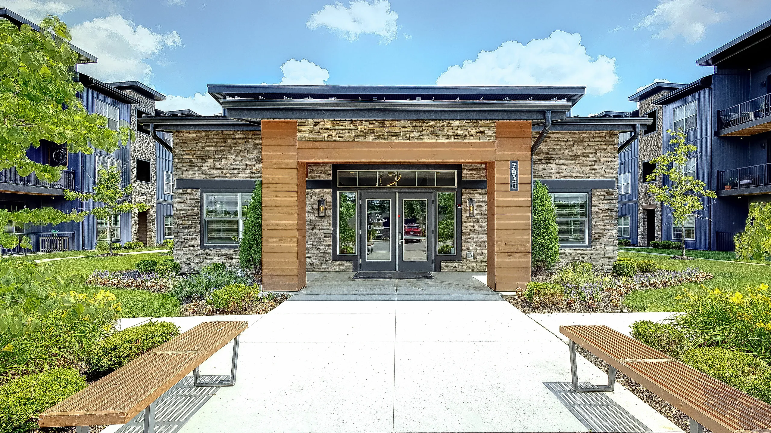 Inviting modern apartment complex entrance with lush landscaping, stone and wood accents, and welcoming outdoor seating under a bright blue sky.