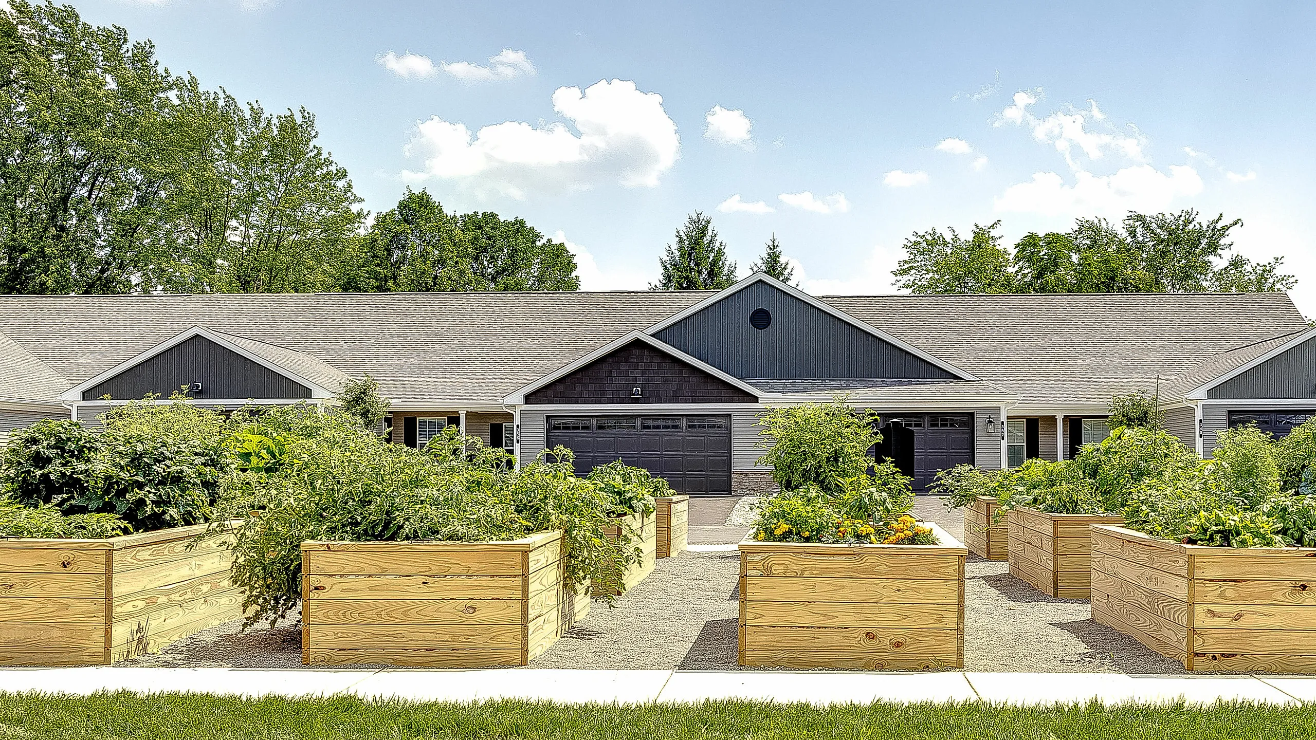 Charming single-story duplex with stylish gray facade and vibrant raised garden beds, framed by lush green trees under a clear blue sky.