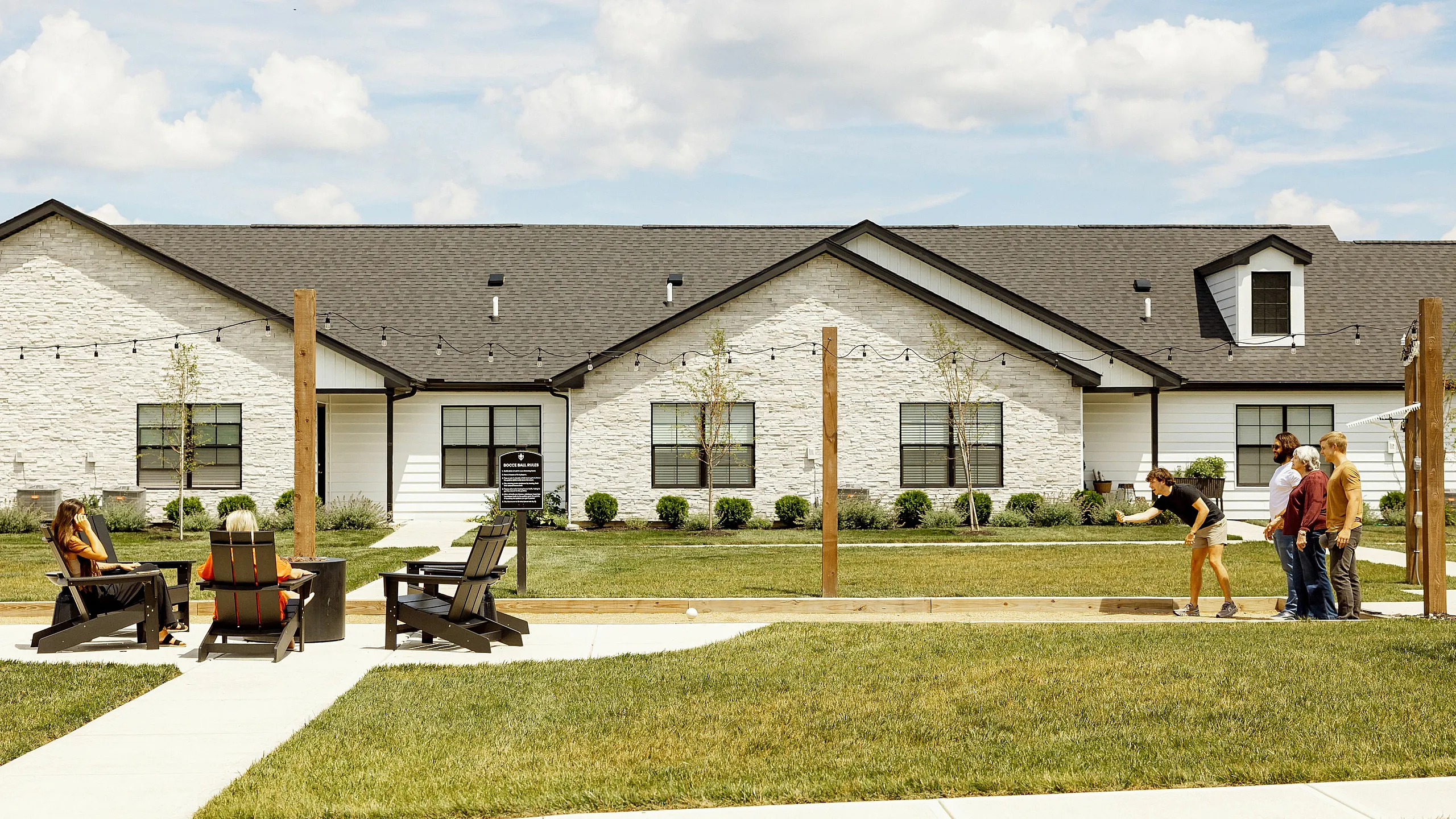 Charming residential community featuring a beautifully manicured lawn, outdoor seating area, and friendly residents enjoying a game under a blue sky.