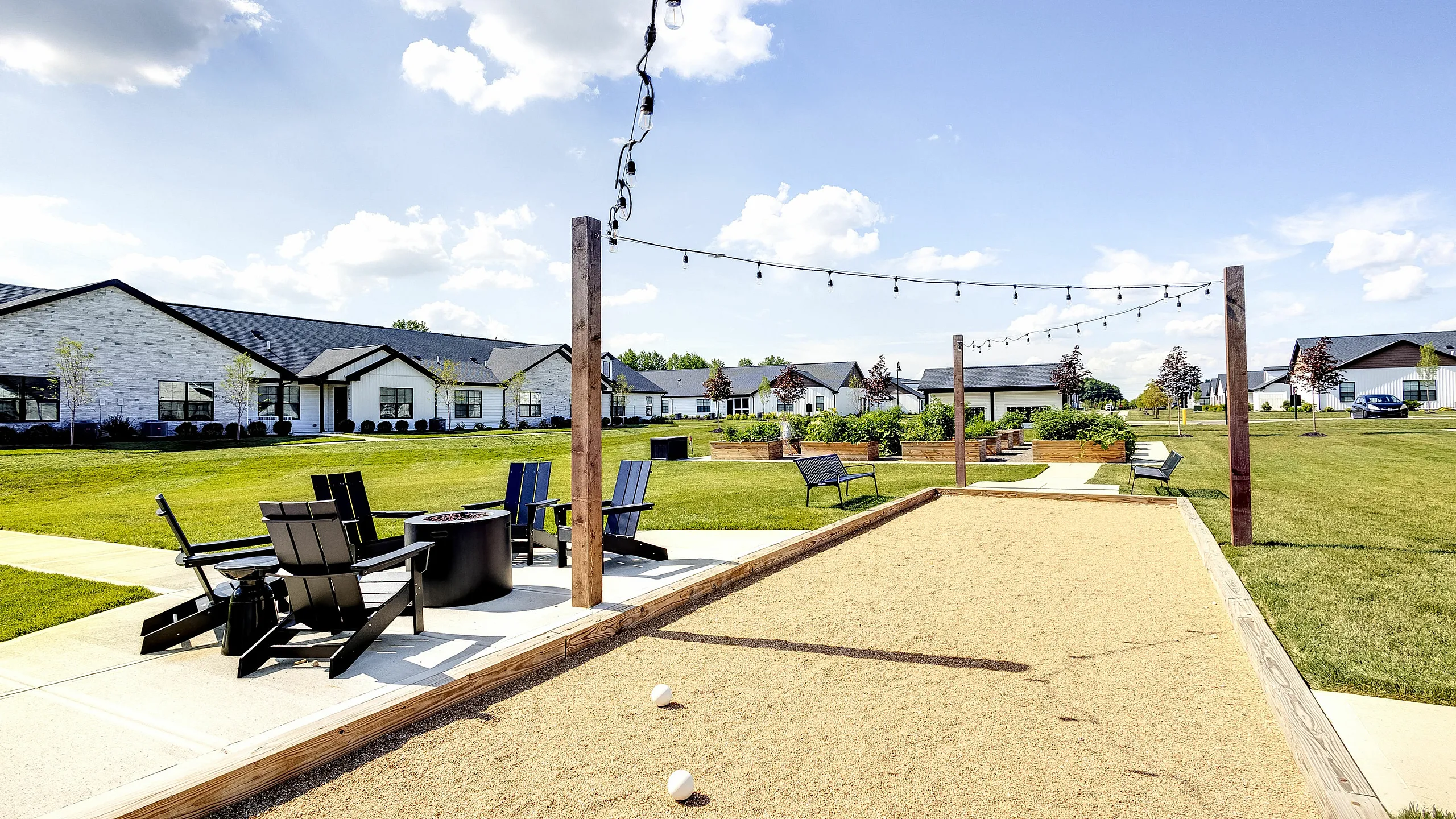 Inviting outdoor recreation area with bocce court, cozy seating around a fire pit, and lush green surroundings under a bright blue sky.