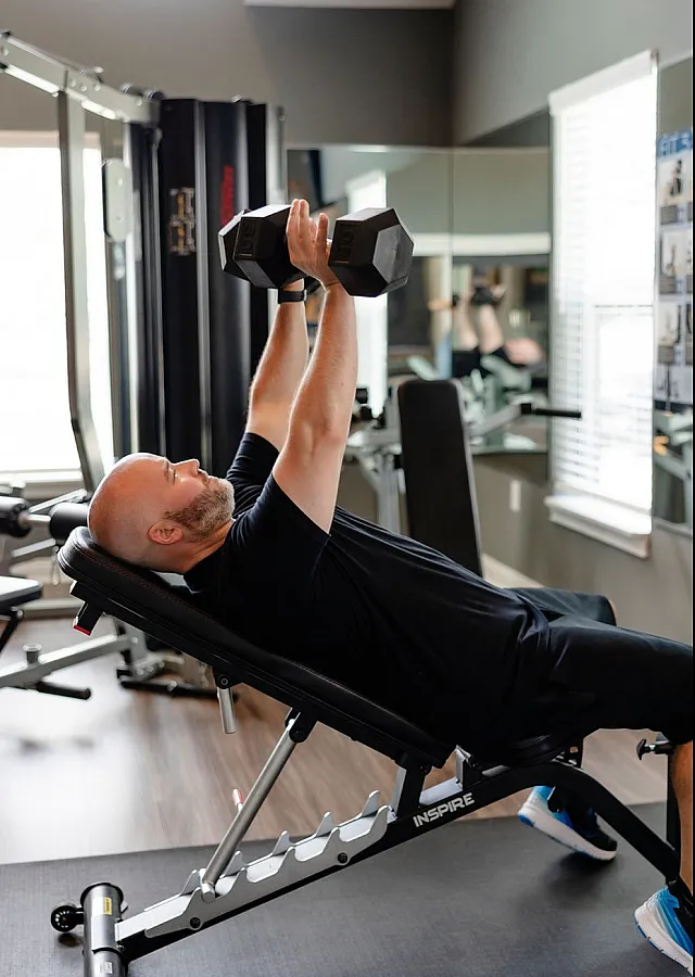 Modern home gym featuring a person lifting dumbbells on an incline bench, surrounded by sleek equipment and large windows for natural light.
