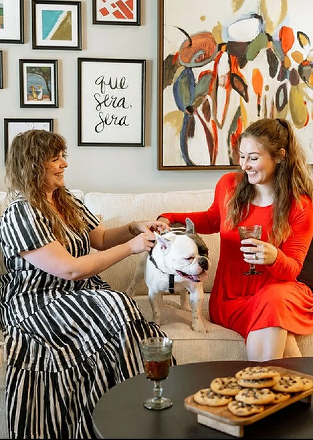 Cozy apartment living room with stylish decor, featuring two women enjoying leisure time with a playful dog on a comfortable sofa amidst vibrant artwork and modern furnishings.