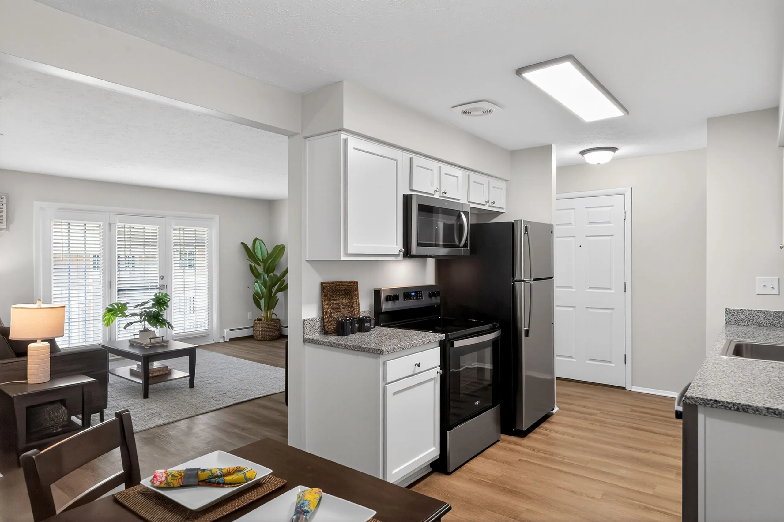 Modern open-plan kitchen and living area featuring sleek stainless steel appliances, elegant white cabinetry, and abundant natural light through large windows with blinds.