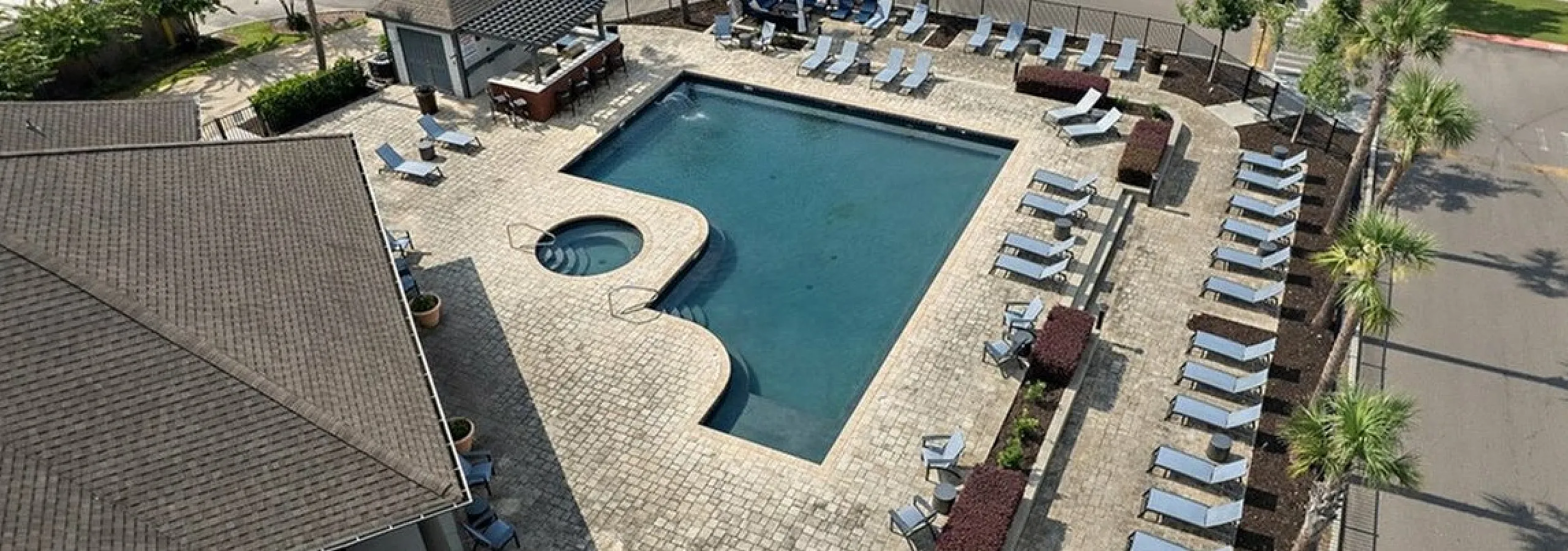 Aerial view of a luxurious apartment complex pool area featuring a sparkling swimming pool, cozy jacuzzi, and numerous sun loungers surrounded by lush palm trees.