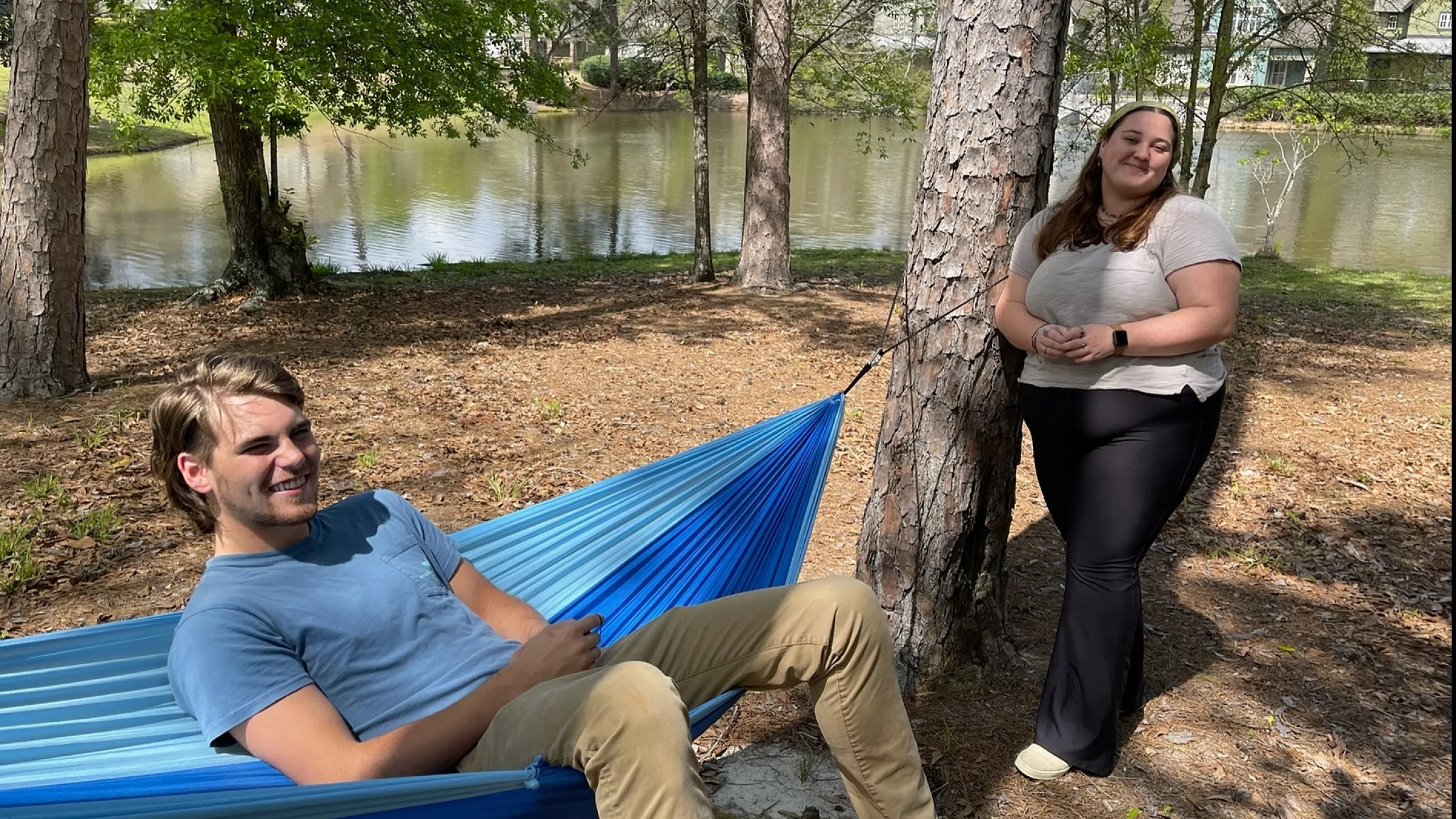 Relaxing outdoor scene with individuals enjoying a peaceful hammock by a serene pond, surrounded by tall trees and natural beauty.