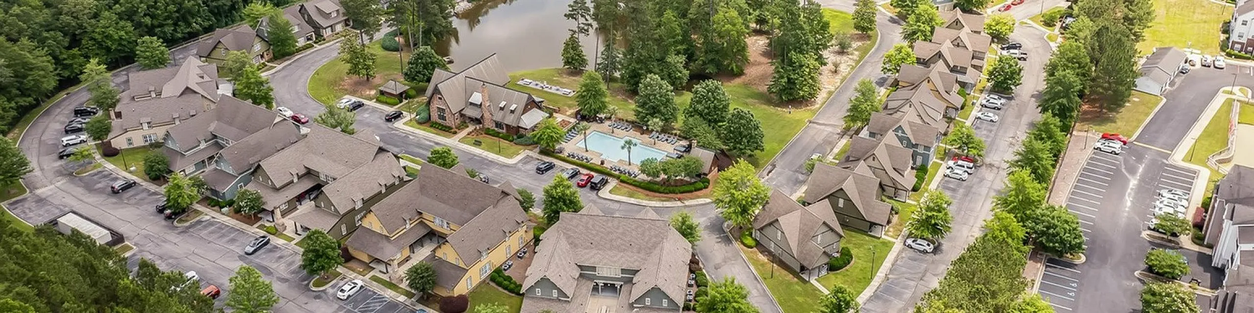 Charming aerial view of a serene residential neighborhood surrounded by lush greenery, featuring a picturesque pond and a large swimming pool.