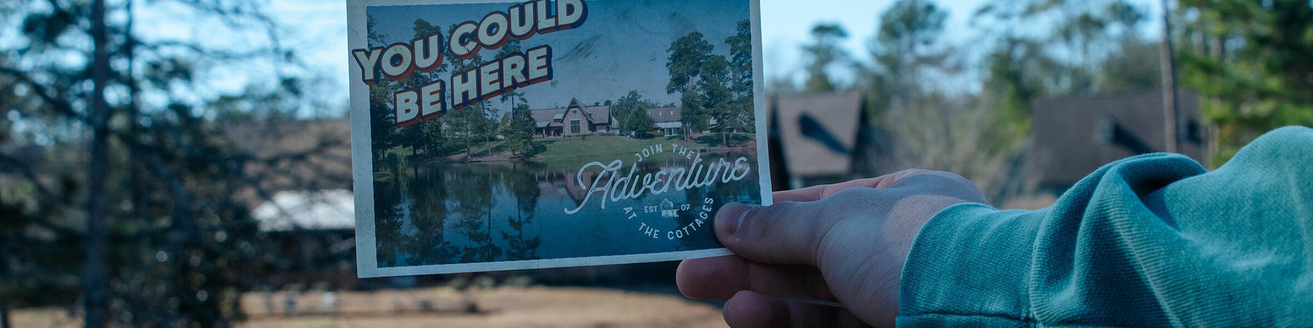 Inviting postcard held by a hand overlooking a serene lake with trees and buildings, suggesting a tranquil getaway destination.