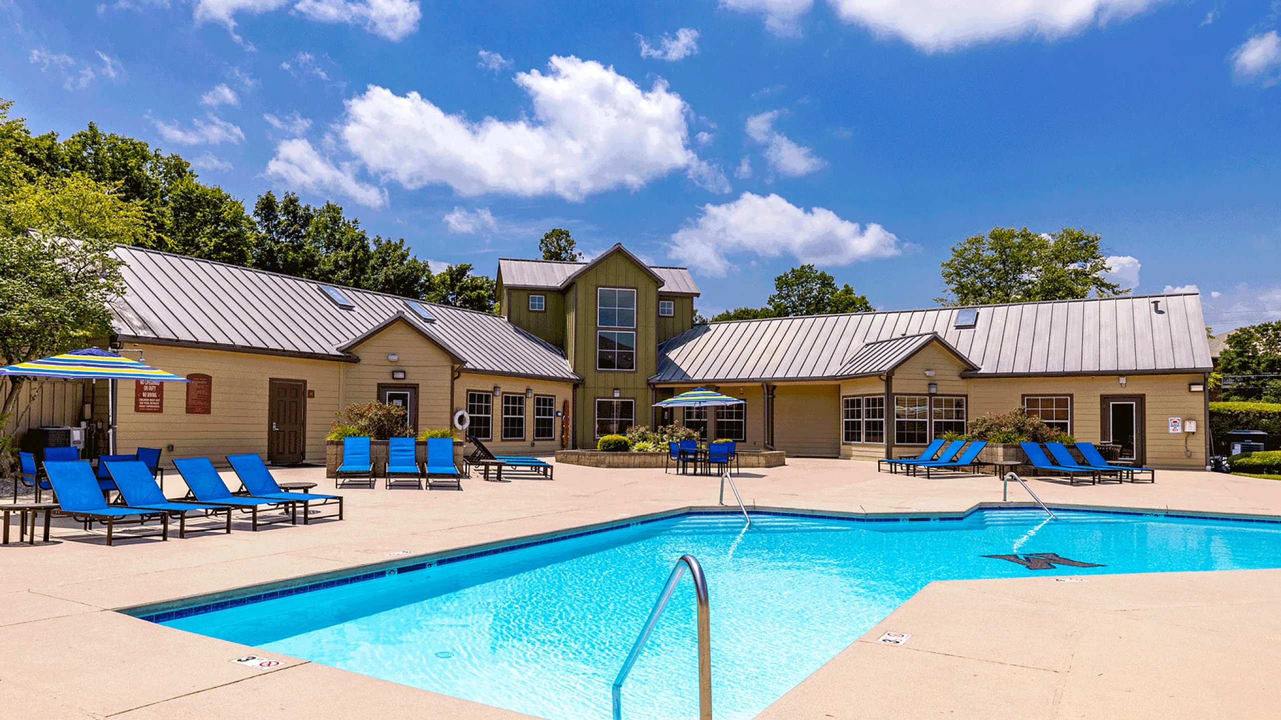 Inviting outdoor pool area with vibrant blue lounge chairs and umbrellas, surrounded by beautiful greenery and a charming clubhouse under a clear blue sky.