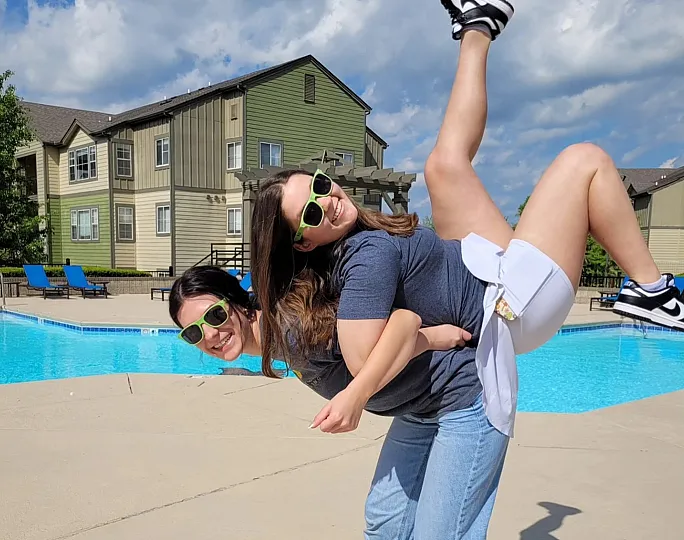 Vibrant community pool area with two people joyfully posing under a bright blue sky, showcasing modern apartment living with lush greenery.