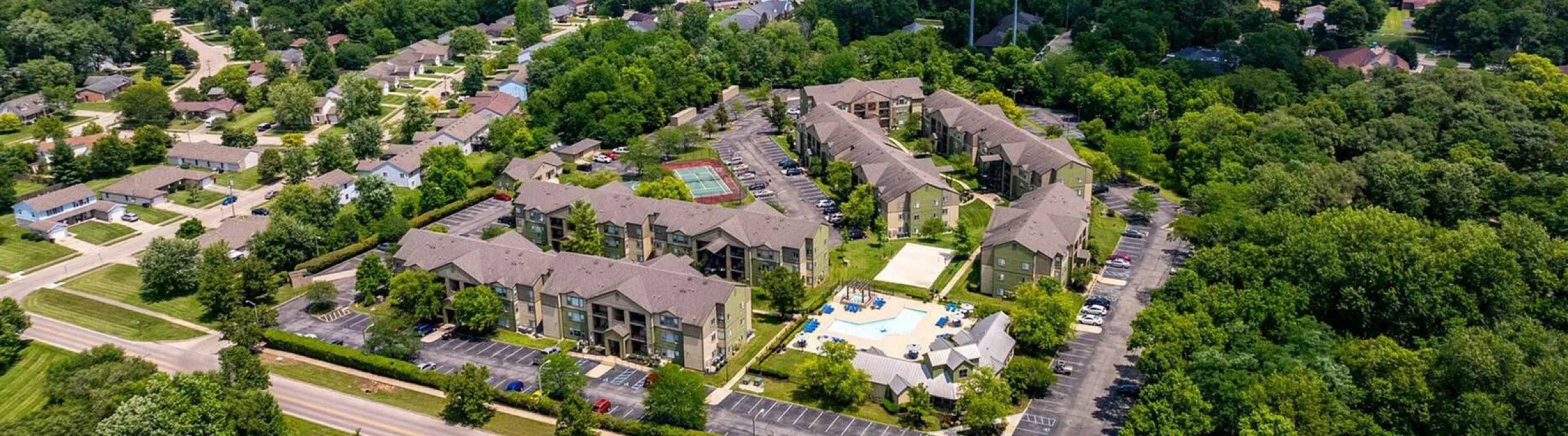 Aerial view of a lush residential complex featuring charming multi-story apartment buildings, tennis court, and a beautiful pool area surrounded by greenery.