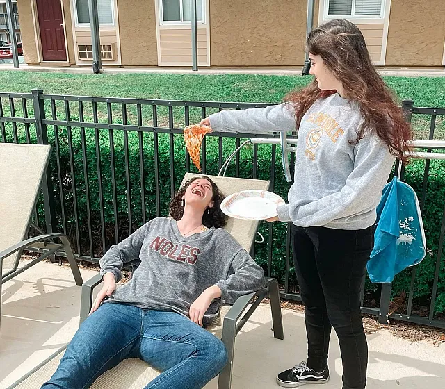 Relaxing outdoor scene with two friends enjoying a playful moment with pizza by a modern apartment complex.