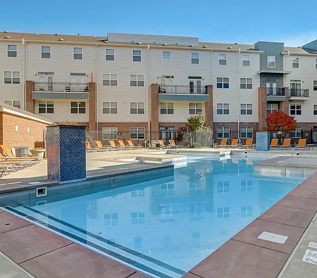 Inviting apartment complex featuring a sparkling swimming pool, serene lounging area, and modern architecture under a clear blue sky.
