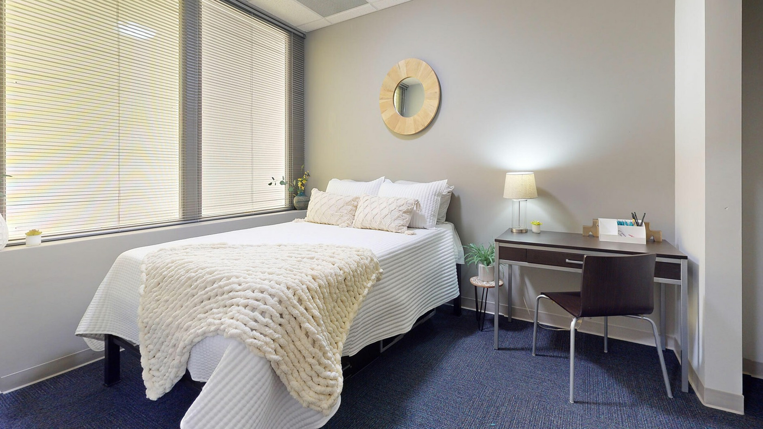 Inviting bedroom with a cozy bed draped in a plush white throw, complemented by a sleek desk and chair, illuminated by a stylish lamp. Large windows allow ample natural light, enhancing the serene and elegant ambiance.