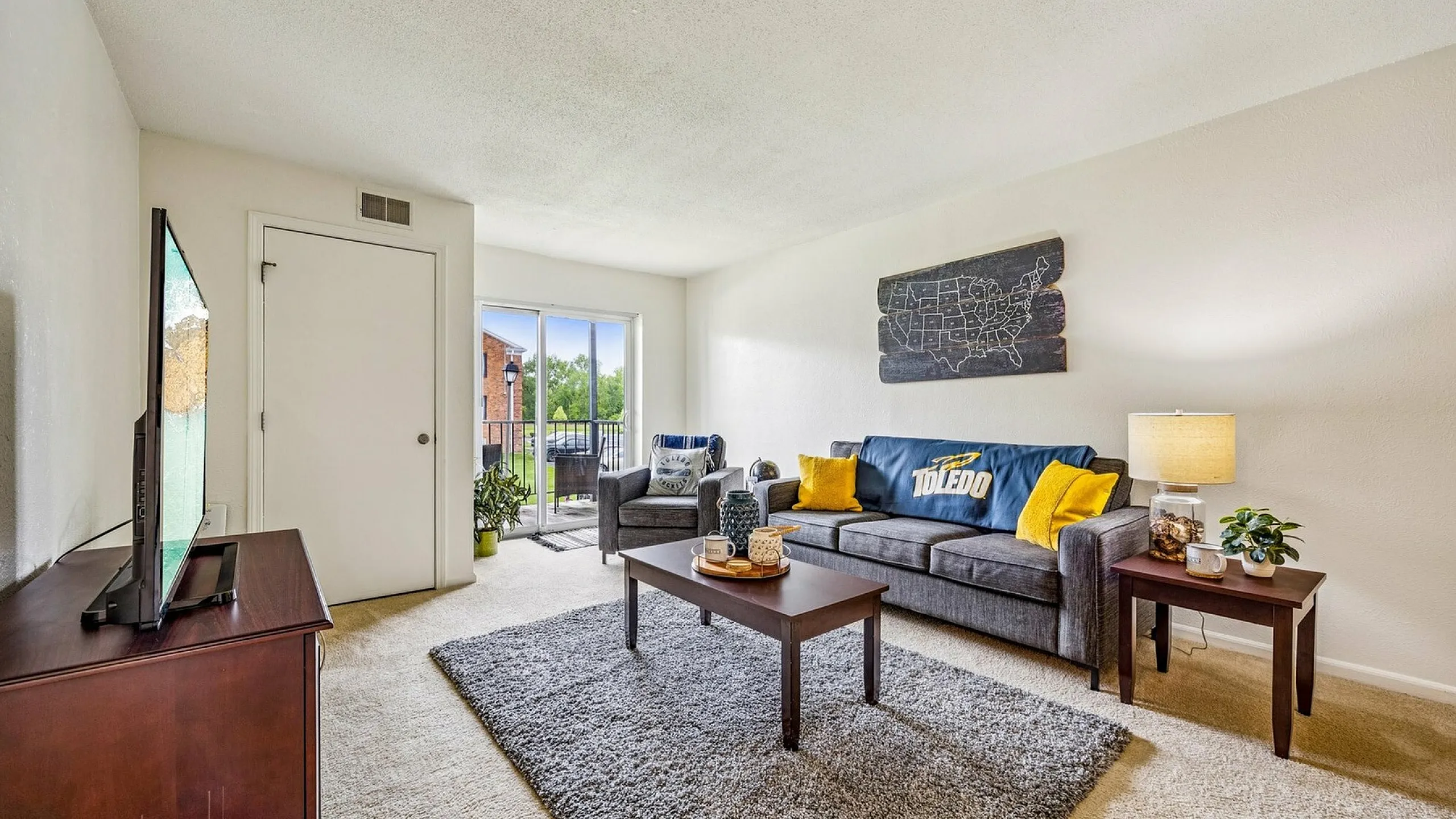 Bright living room with cozy seating, large window doors opening to a balcony, plush gray carpet, and stylish decor in neutral tones.