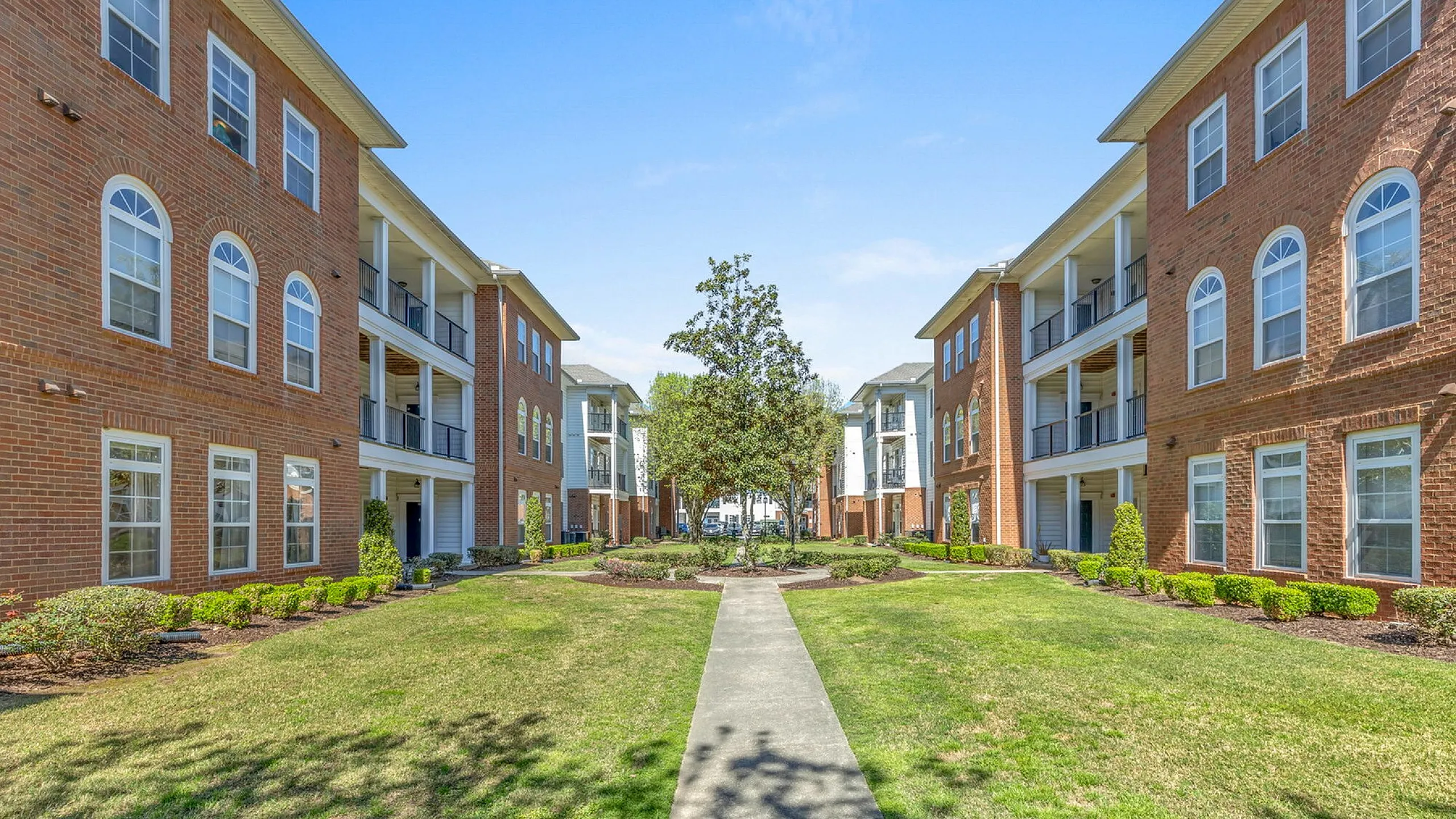 Charming brick apartment buildings with elegant balconies overlooking a lush, landscaped courtyard path under a clear blue sky.