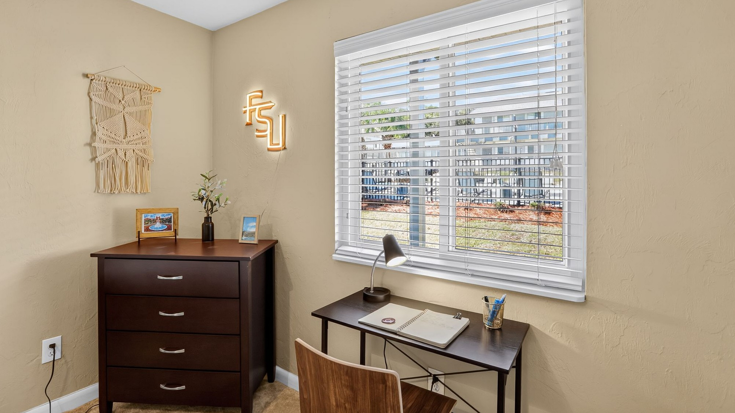 Bright home office space featuring a modern wooden desk with a lamp, a chic macrame wall hanging, and a window with a pleasant garden view.