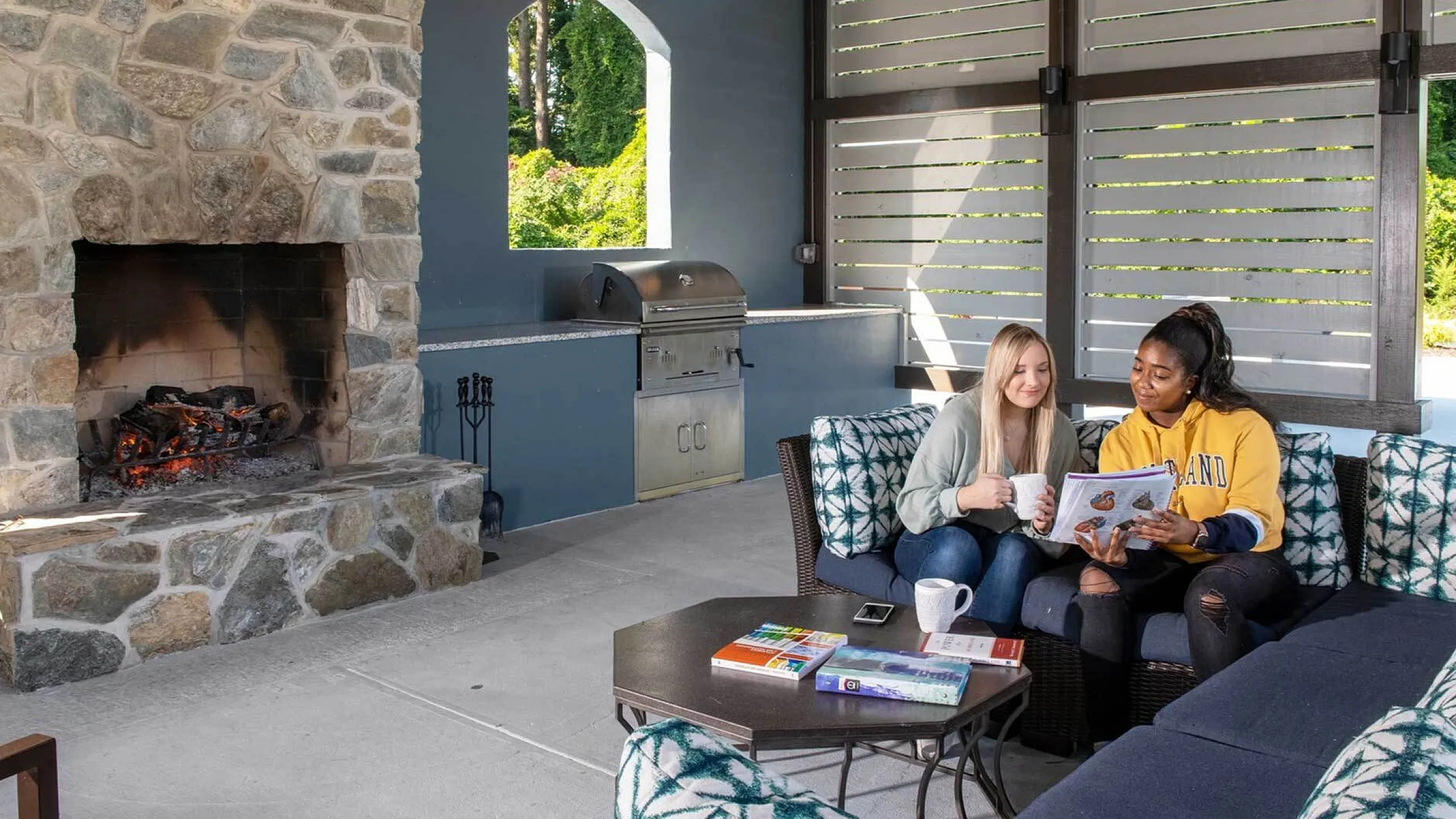 Charming outdoor seating area with a cozy stone fireplace, featuring two people enjoying a relaxed moment with books and coffee.