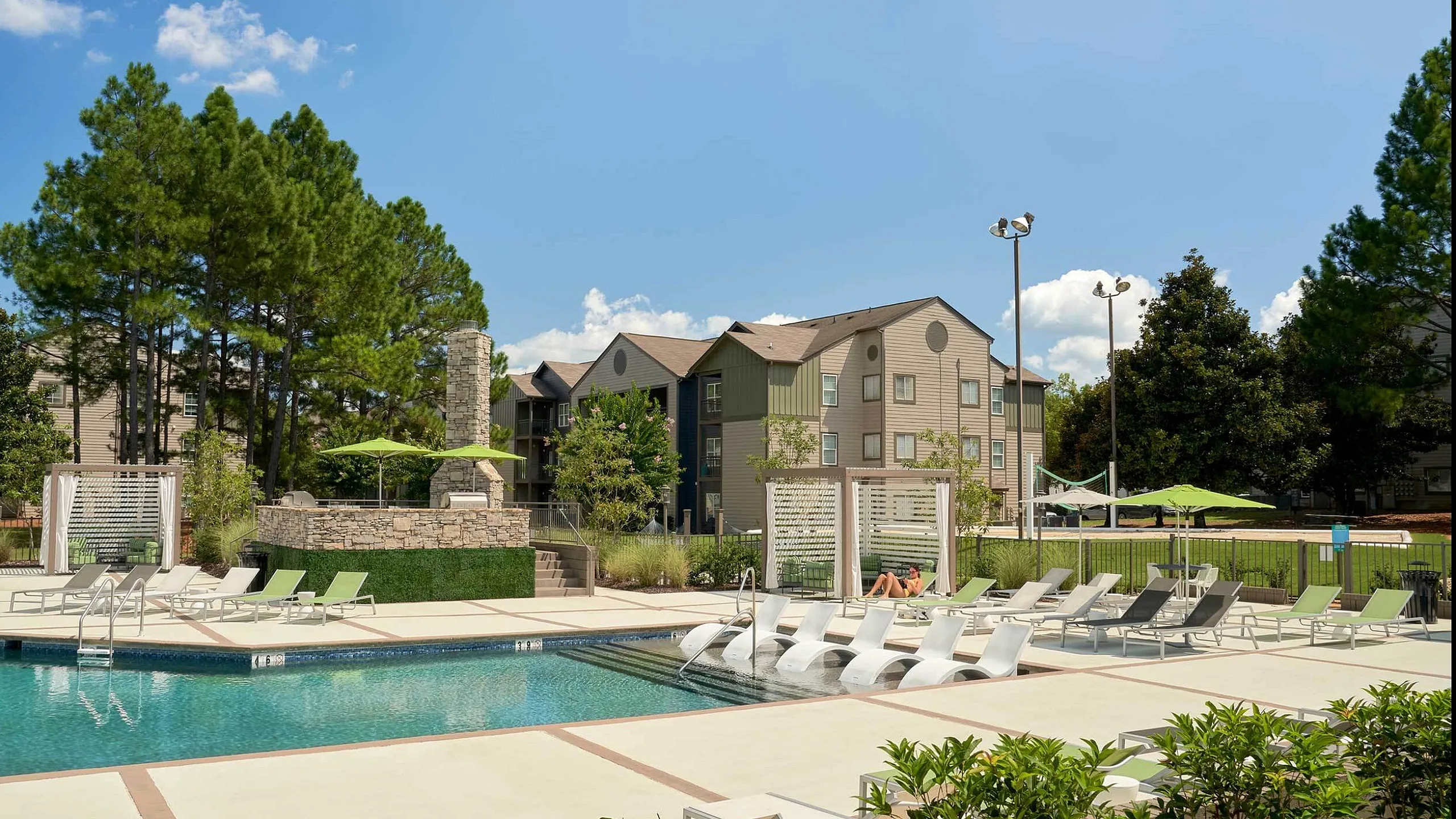 Luxurious apartment complex with a sparkling pool, relaxing lounge chairs, and lush greenery under a clear blue sky.
