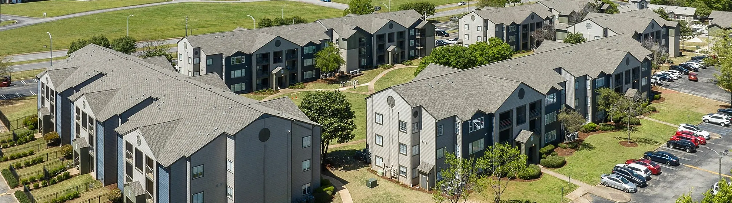 Aerial view of a modern apartment complex with multiple buildings, surrounded by lush greenery and ample parking, set in a serene suburban landscape.