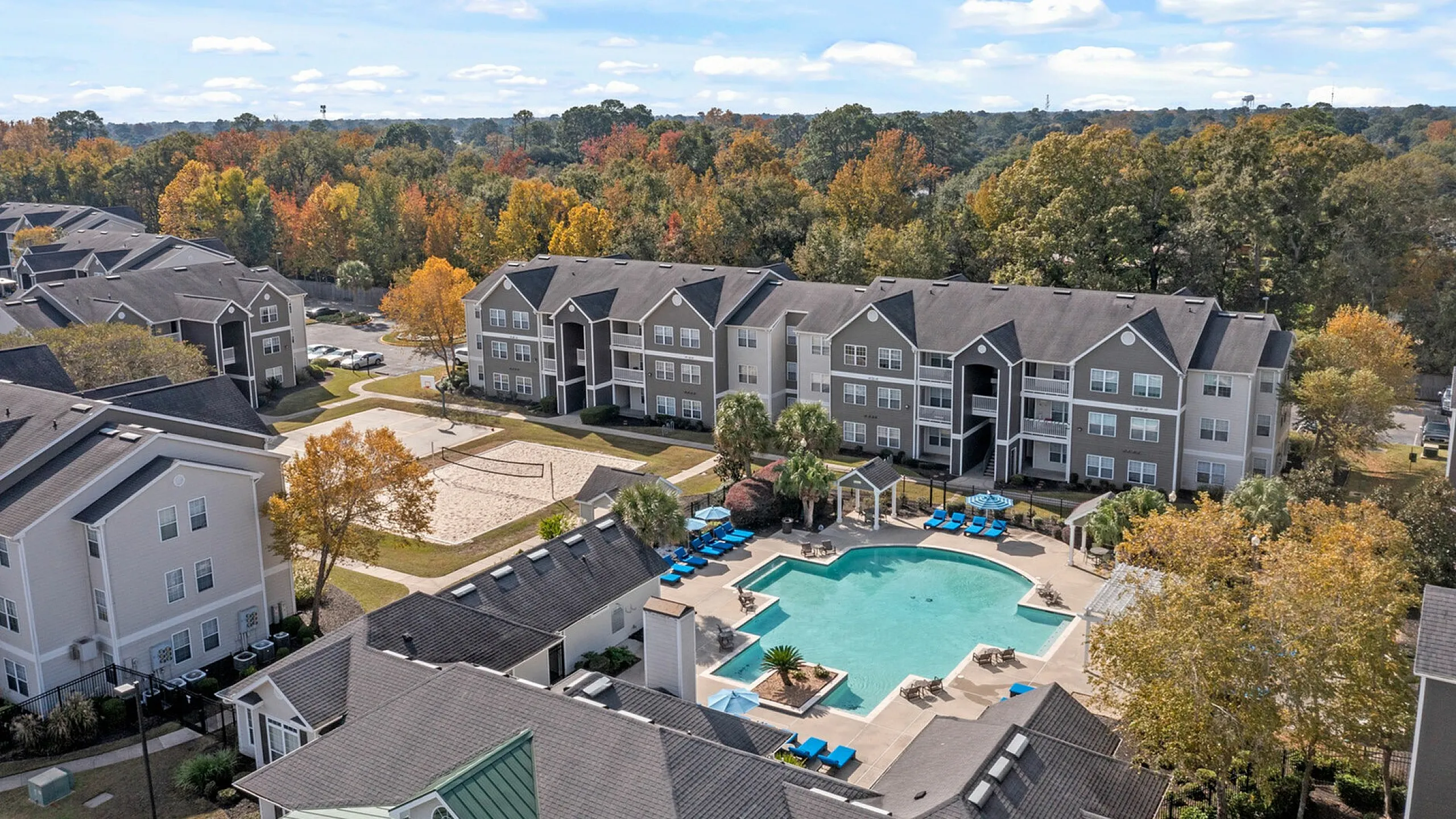 Beautiful aerial view of modern apartment complex with a sparkling pool, surrounded by lush autumn trees and spacious recreational areas.
