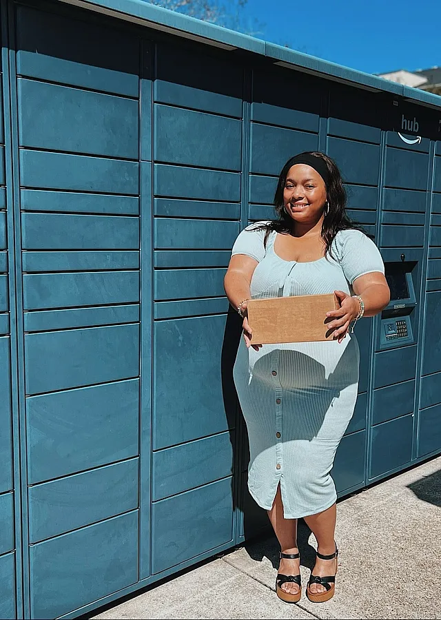 Smiling woman in a light dress holding a package in front of a row of blue parcel lockers under a clear sky.
