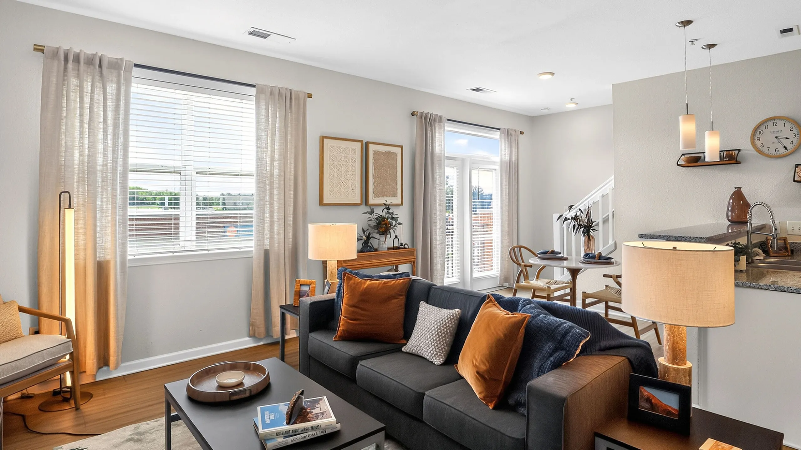 Inviting living room with modern decor, featuring a comfy dark sofa adorned with orange pillows, elegant curtains, and abundant natural light. Open layout includes a stylish dining area adjacent to a beautifully designed kitchen.