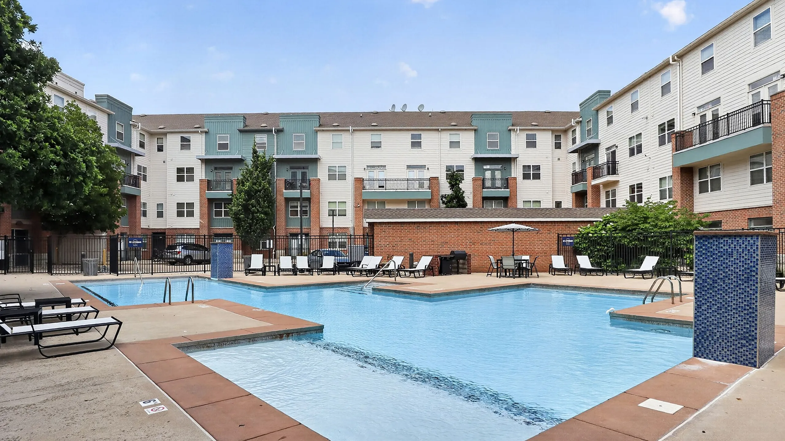 Inviting apartment complex featuring a spacious pool area with modern lounge chairs, vibrant landscaping, and multi-story residential buildings in the background.
