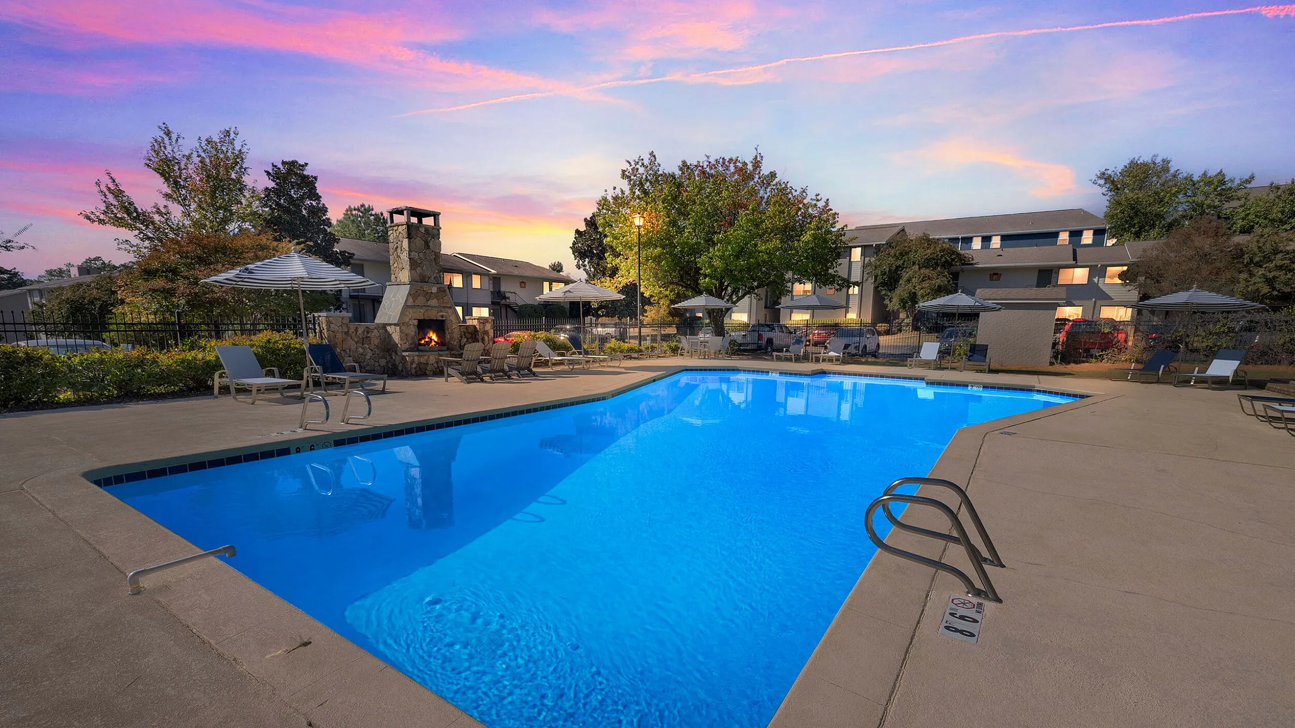 Inviting community pool area at sunset with cozy lounge chairs, umbrellas, and a charming stone fireplace, perfect for relaxation.