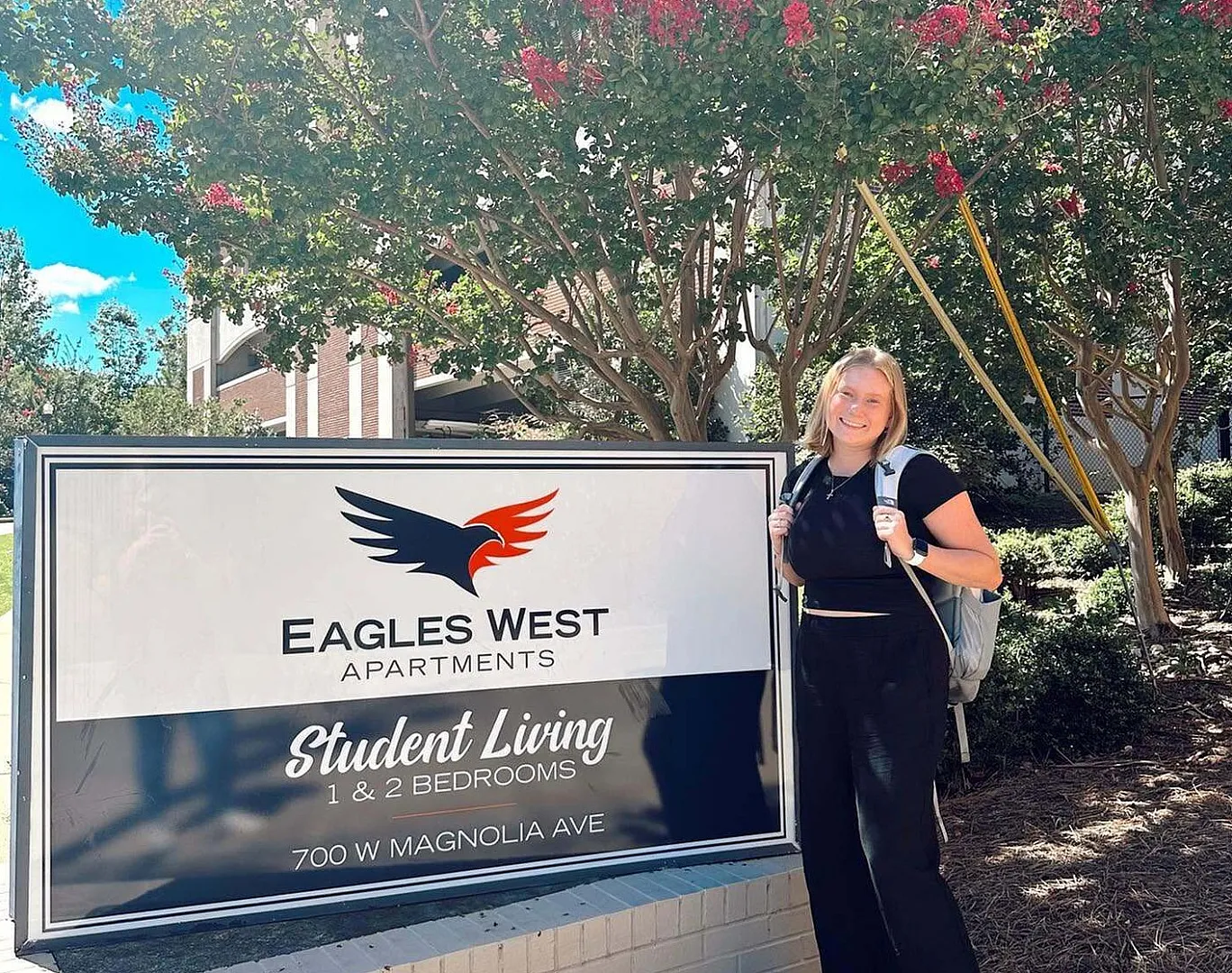 Eagles West Apartments sign for student living featuring 1 and 2 bedroom options, with a person smiling in a vibrant outdoor setting.