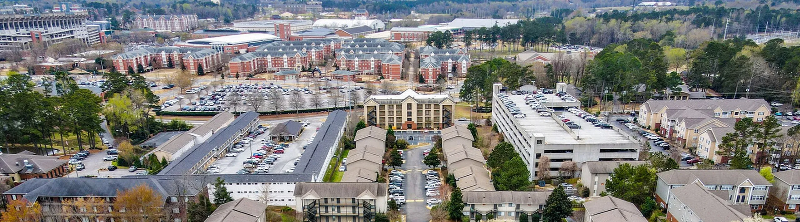 Aerial view of a vibrant apartment complex featuring modern residences, ample parking, lush landscaping, and a sparkling central pool, set against a backdrop of a bustling city and green parks.