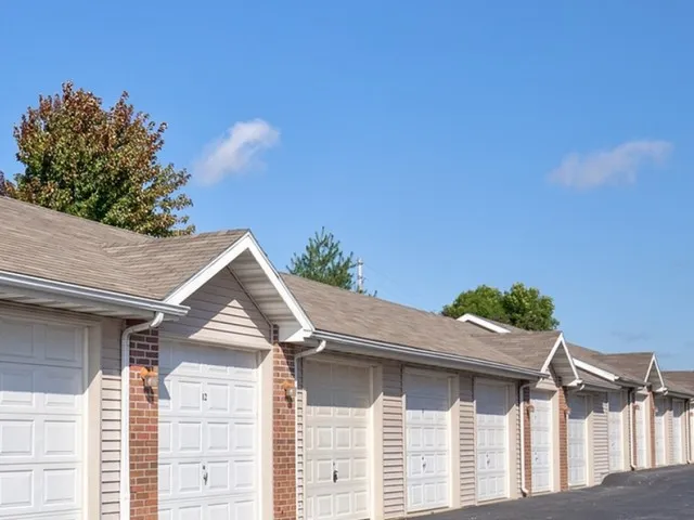 Exterior view of a row of light-gray garage units with white roll-up doors and pitched roofs under a clear blue sky at Embassy Apartments in Ozark, Missouri.