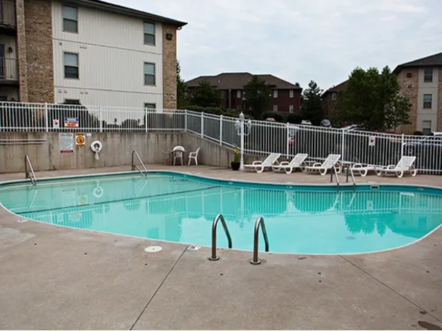 Outdoor swimming pool with clear water, surrounded by lounge chairs, a safety fence, and adjacent apartment buildings.
