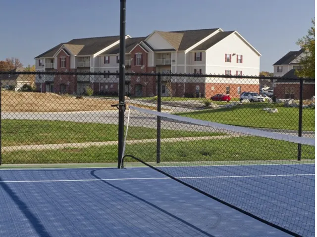 Outdoor tennis court with a blue surface surrounded by a black chain-link fence. In the background, a residential building and clear blue sky.