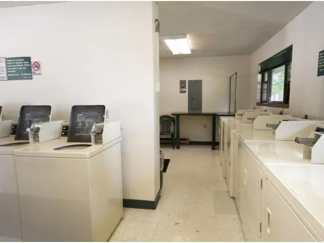 Community laundry facility with multiple top-loading washers and dryers, bright lighting, and a folding table near the back wall.